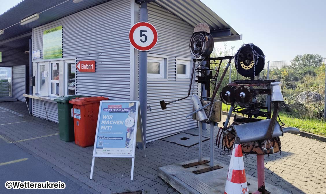 Ein Wertstoffhof mit zwei Schrott-Statuen, neben denen ein "Wir machen mit!" Plakat aufgestellt ist.